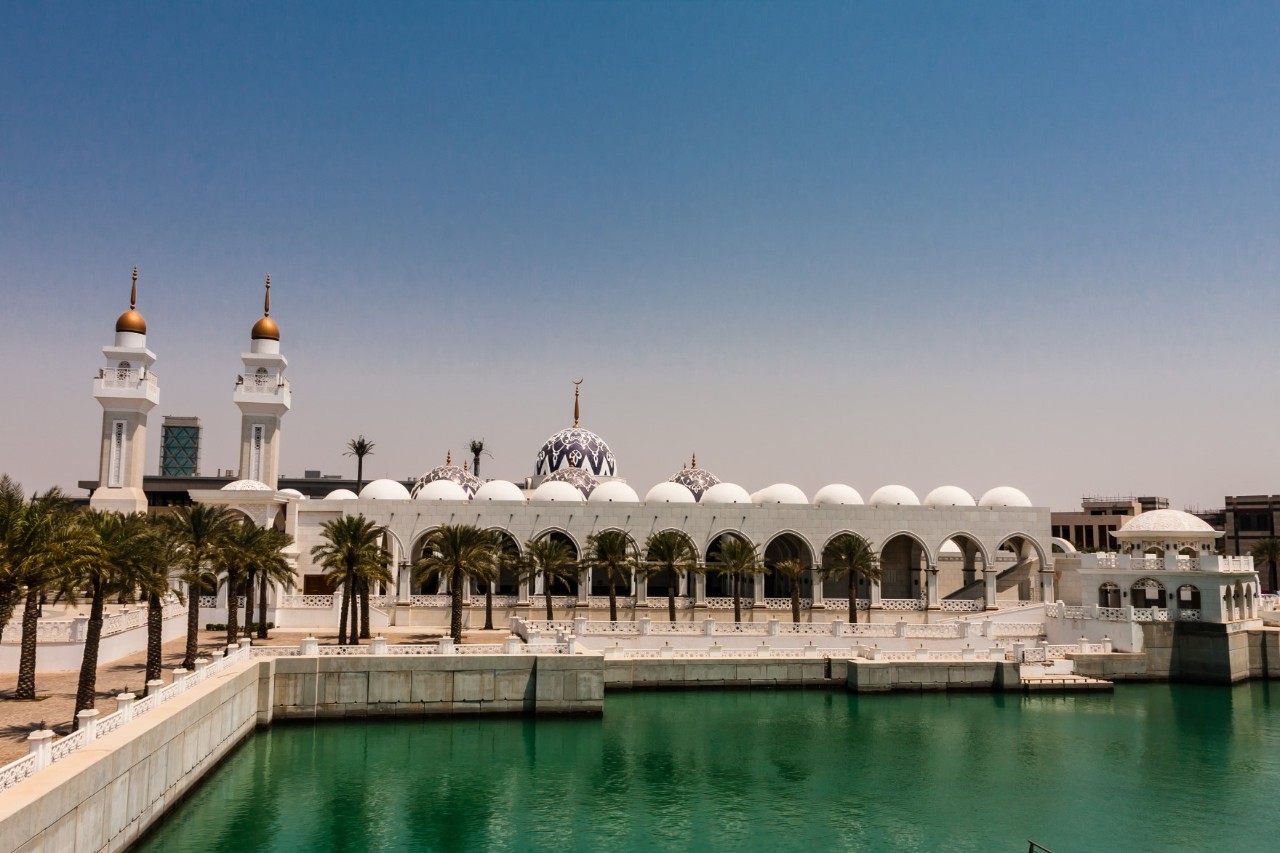 A white mosque with minarets, in front a water basin with a concrete wall surrounded by palm trees. 