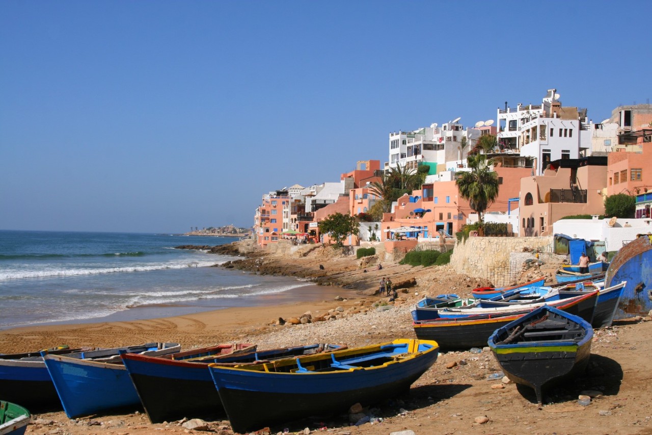 A coastal village with reddish and white houses stretching directly along the sea on a small hill. Several small blue fishing boats lie in the sand in the foreground.