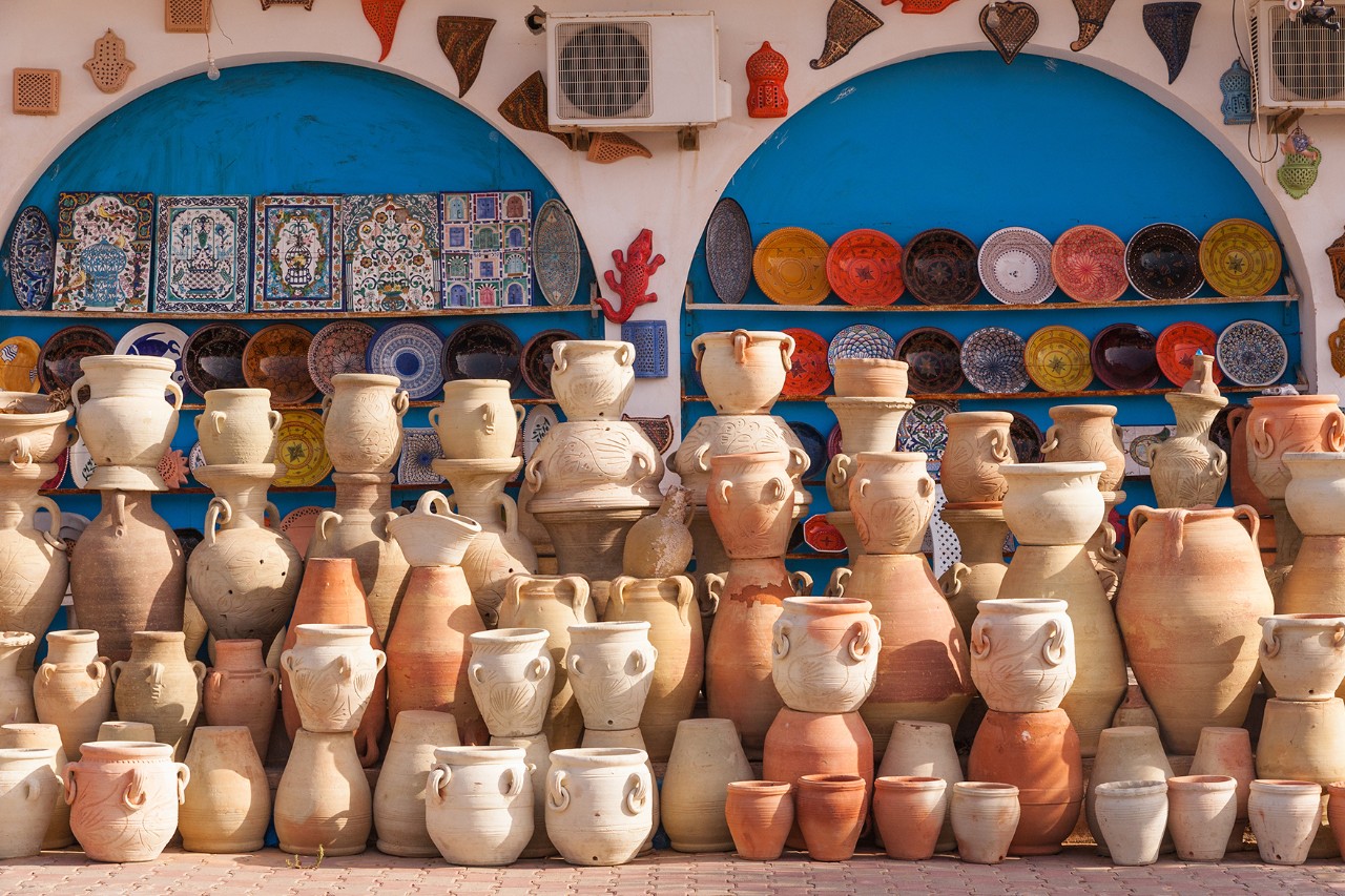 Handmade pottery in various shapes and sizes. In the background are painted ceramic plates and decorated tiles, displayed in two blue wall niches.