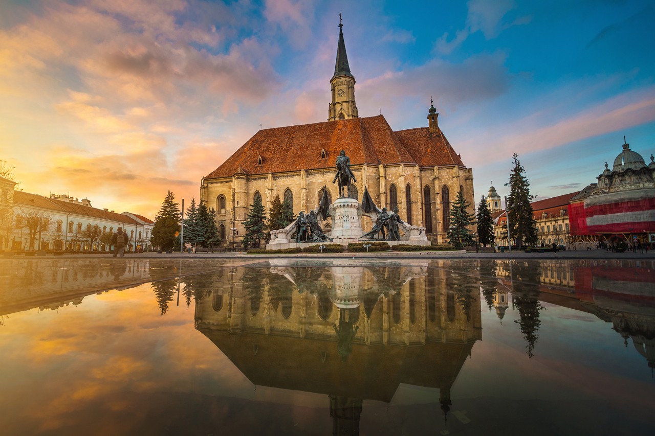 The picture shows a bright church with a large nave and a tall tower. In front of it stands an equestrian statue. The church and statue are reflected in a smooth water surface in the foreground.