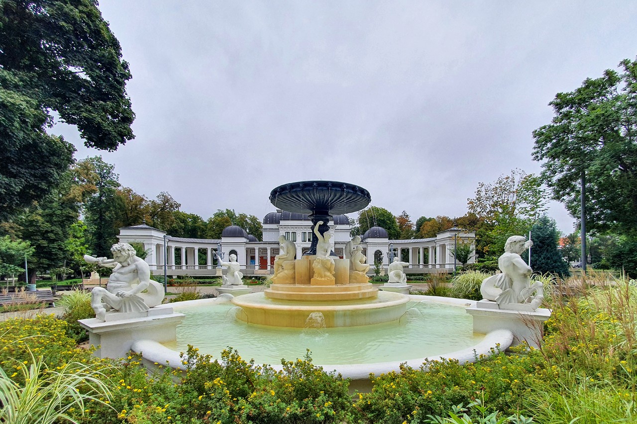 In the foreground is a fountain decorated with statues, surrounded by green trees and shrubs. A colonnade can be seen in the background.