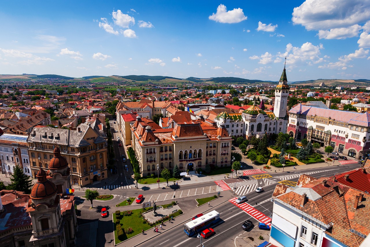 The picture shows a city from above. A domed building and a tall tower stand out in particular. Green hills and a blue sky can be seen in the background.
