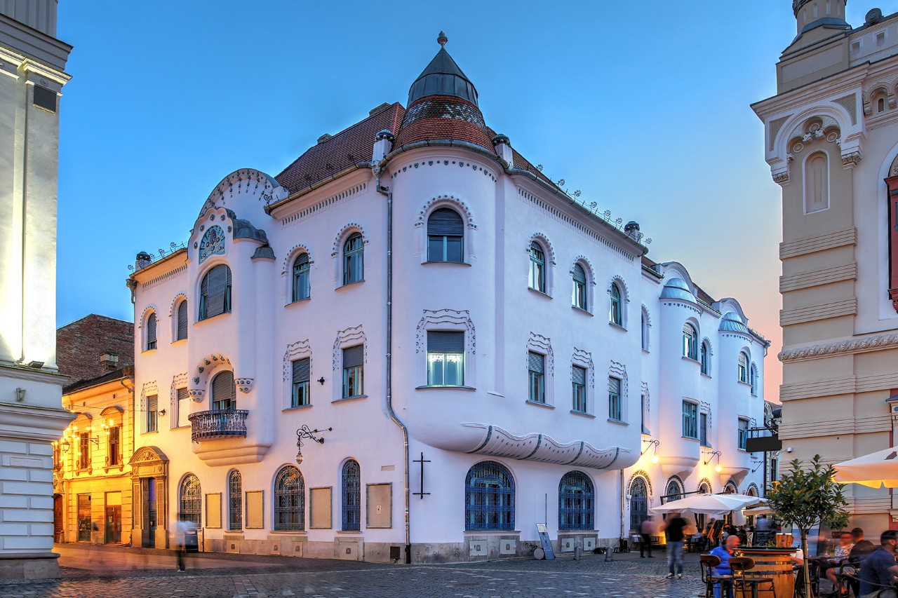 White Art Nouveau building with a curved façade on a street corner. On the right-hand side of the picture is a café with outdoor seating.