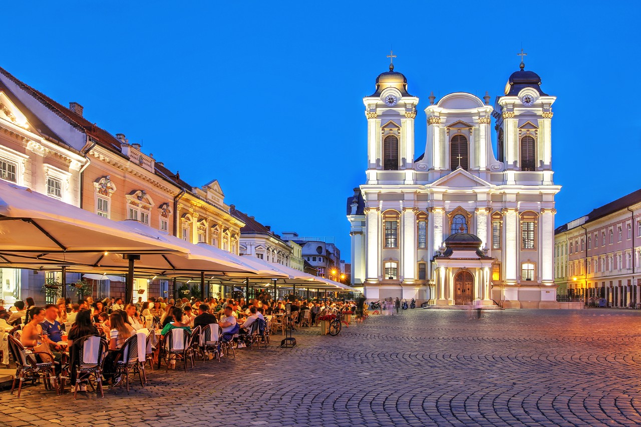 Market square at dusk with lively outdoor dining on the left and a brightly lit church with two towers in the background.