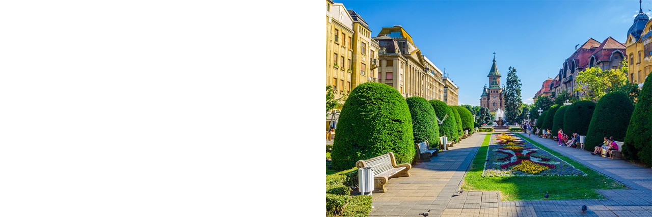 Well-maintained park with round, green hedges, people sitting on benches and a flower bed in the middle, surrounded by historic buildings. A cathedral can be seen in the background.