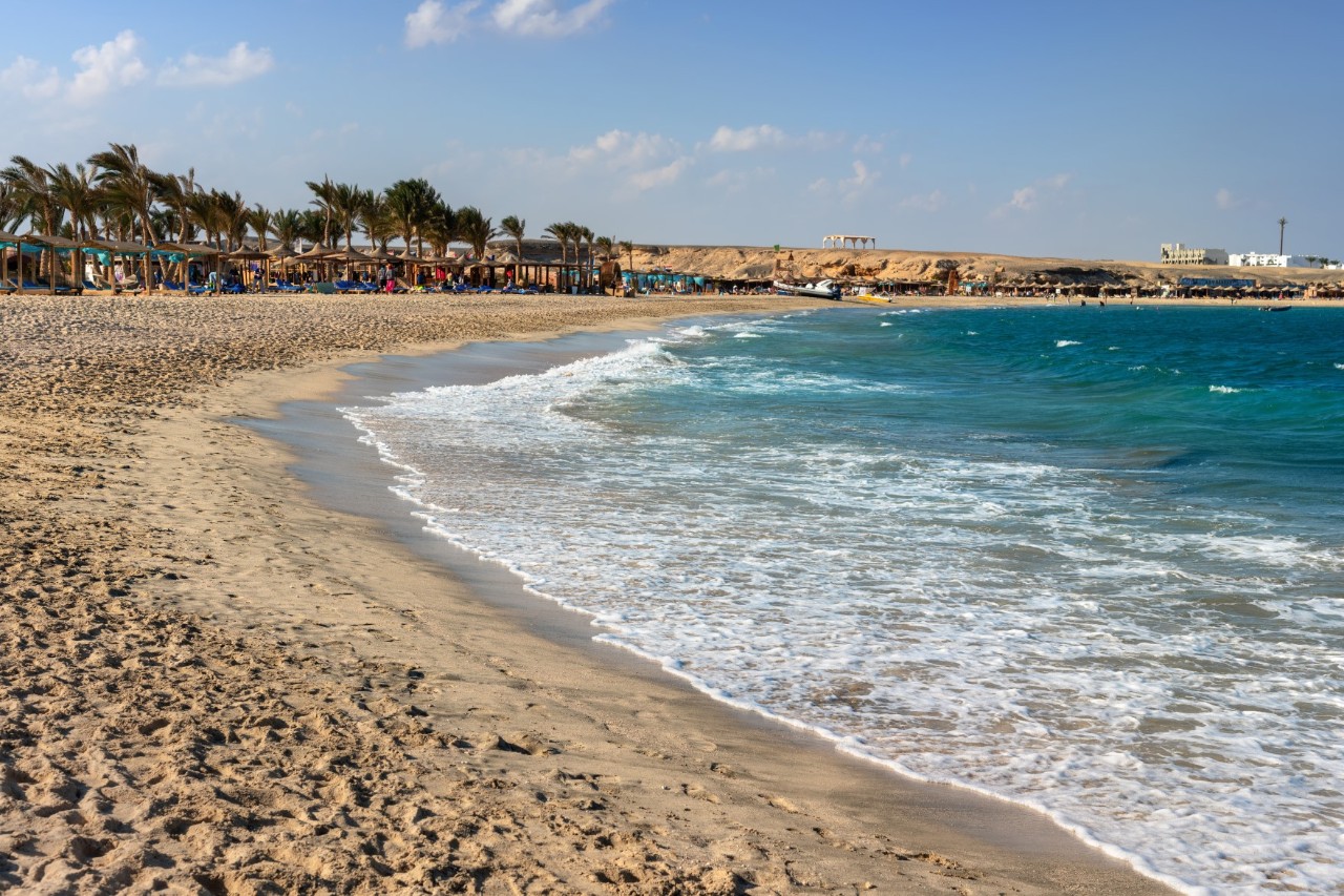 A sandy beach with gentle waves. In the background, there are palm trees, beach huts and sun umbrellas.