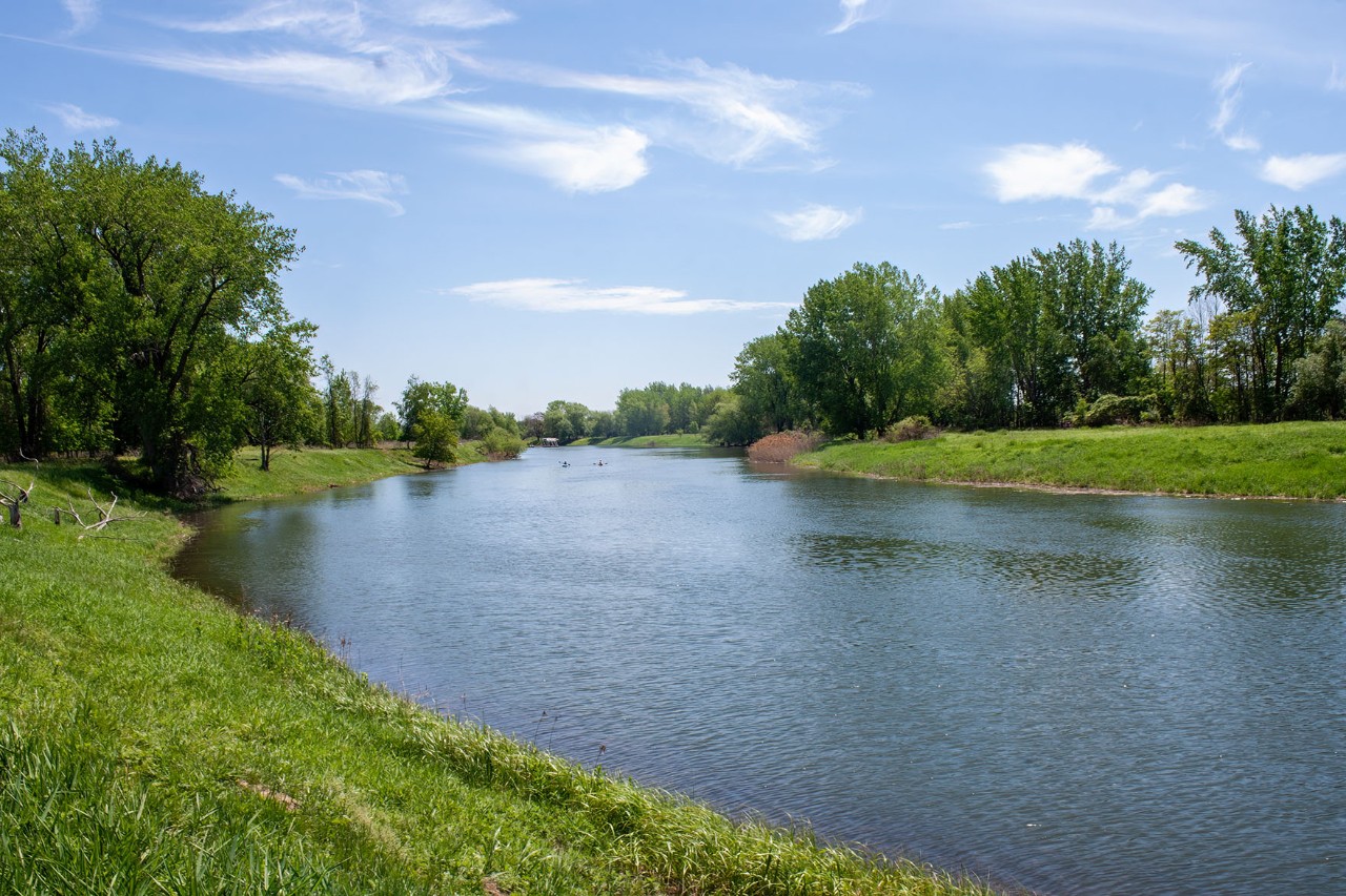 Wide section of a river with green banks and trees under a blue sky with a few clouds, in a park.