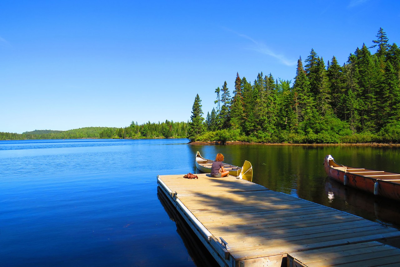 A tranquil lake with deep blue water, surrounded by dense, green forests. In the foreground is a wooden jetty where a person is relaxing in the sun. Next to it are canoes in the water.  