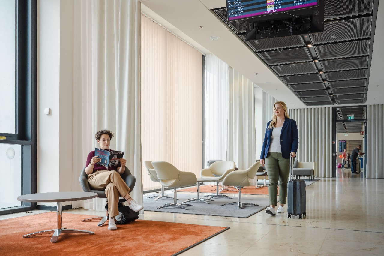 Eine Frau sitzt an einem Tisch und liest ein Magazin, eine weitere Frau geht den Gang mit einem Koffer entlang. ©Ekaterina Zershchikova / Flughafen Berlin Brandenburg GmbH