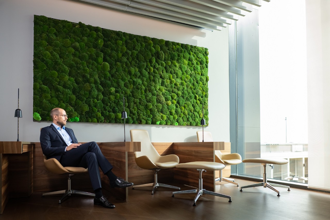A man in a suit, around 40 years old, sits in one of several lounge chairs and looks out over the apron. Behind him is a green wall planting.