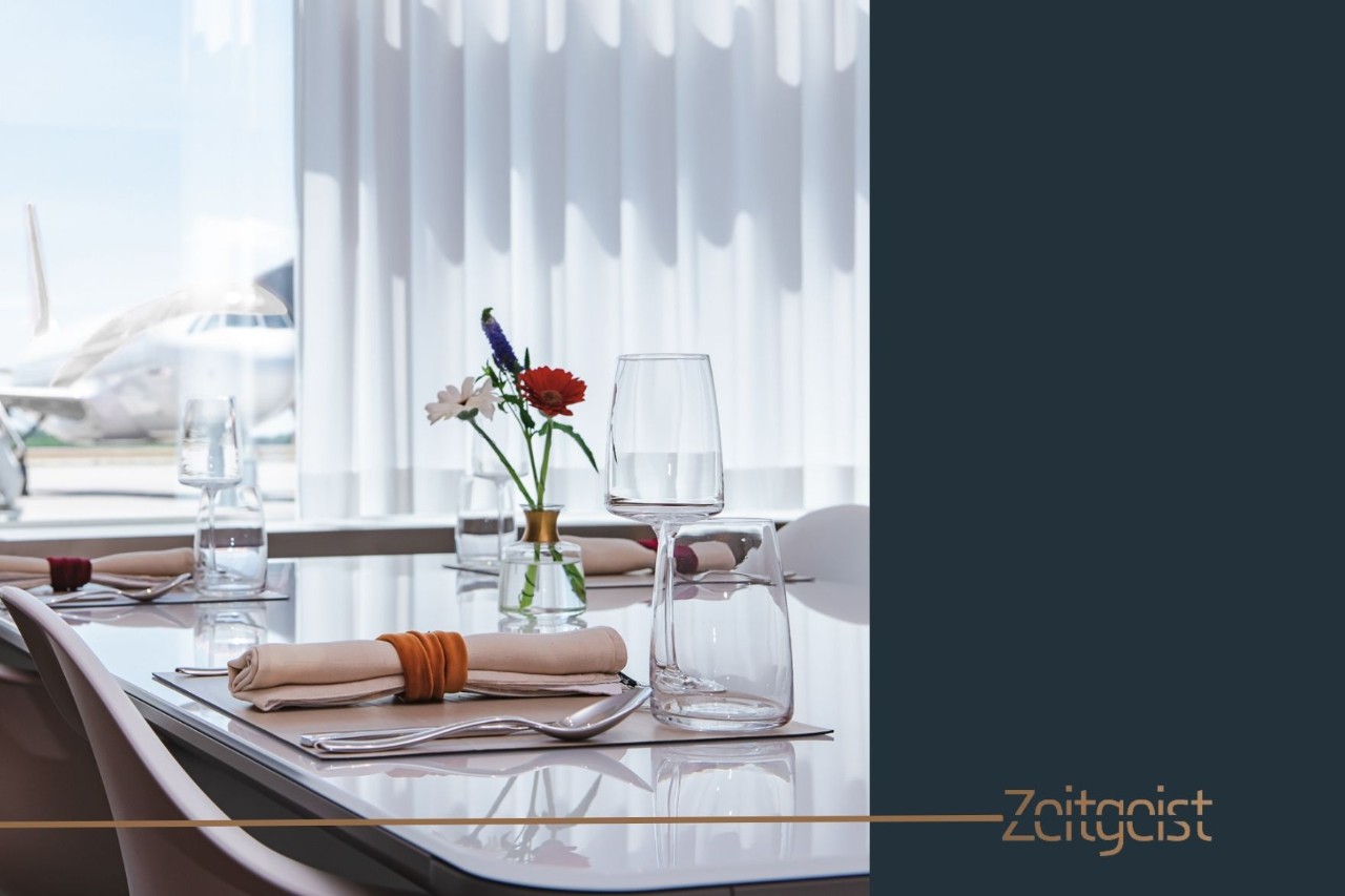 Set table in the VIP area. On the table are glasses, a vase with colourful flowers and cutlery. An aeroplane can be seen through the window in the background.