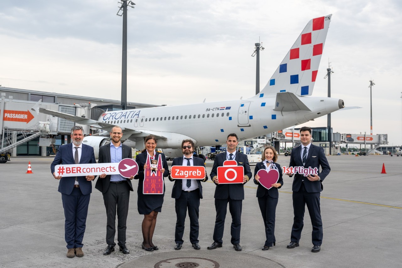 Seven people stand in front of a Croatia Airlines plane. They are holding up various signs to mark the airline's maiden flight from BER to Zagreb.