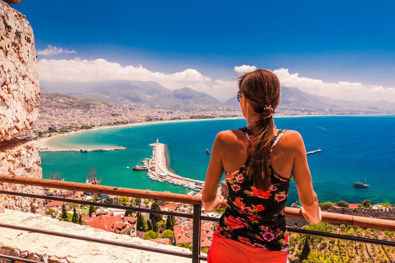 View from behind of a woman in light summer clothing on a hot, sunny summer’s day. She is looking from behind a railing of a light-coloured building over a Mediterranean city to the turquoise waters of the Mediterranean. 