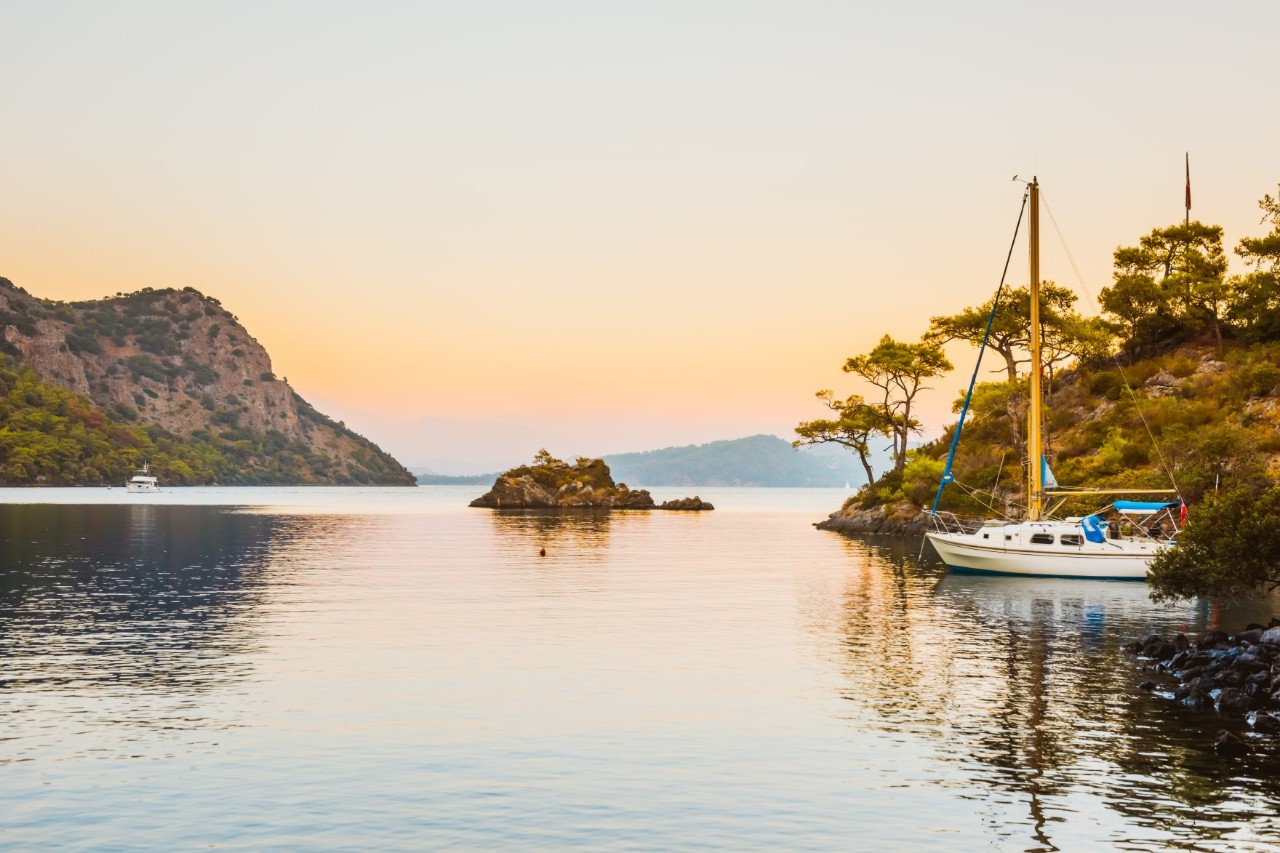View over the water in the evening. On the right, a small sailing boat is anchored in a bay, the surrounding land of the bay is covered with plants and trees. There are mountains to the left of the picture. A small rock is sticking out of the water. 