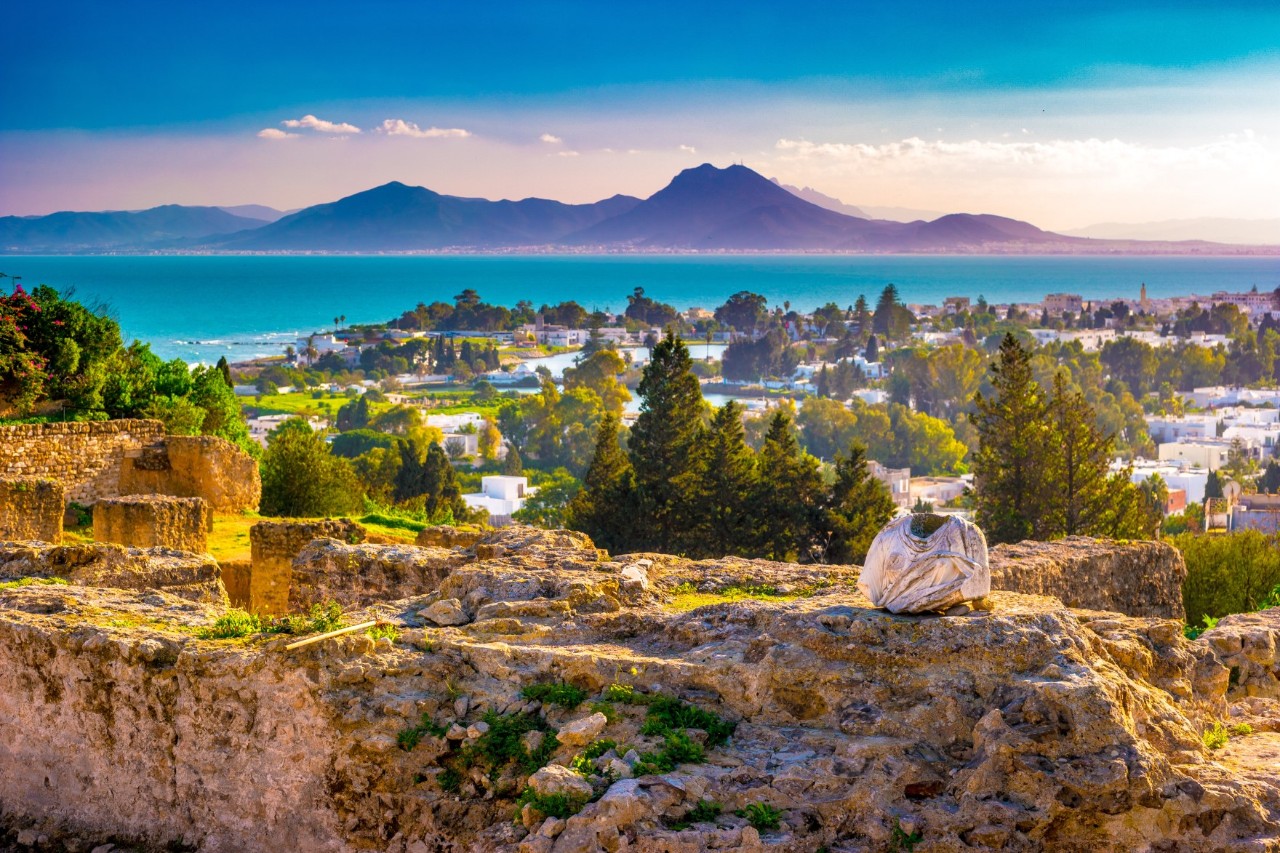 View over ancient stone walls to a city with white buildings and trees in between. Behind the city is the sea with blue water. Mountains behind it again. The sun shines into the picture with evening light.