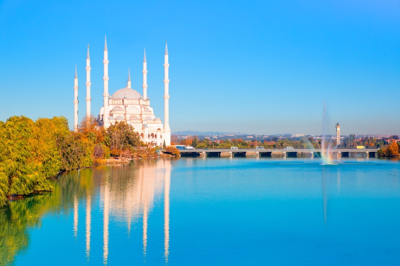 View over a wide river with blue water to a massive stone bridge. A fountain gushes up in front of the bridge, and behind it a city spreads out with a number of buildings with scattered trees. On the left, autumn-coloured trees and, behind the bridge, a large white mosque with six slimline, towering minarets frame the scene. The mosque and the trees are picturesquely reflected in the clear water. The sky is bright blue, and the sun lights up the scene.