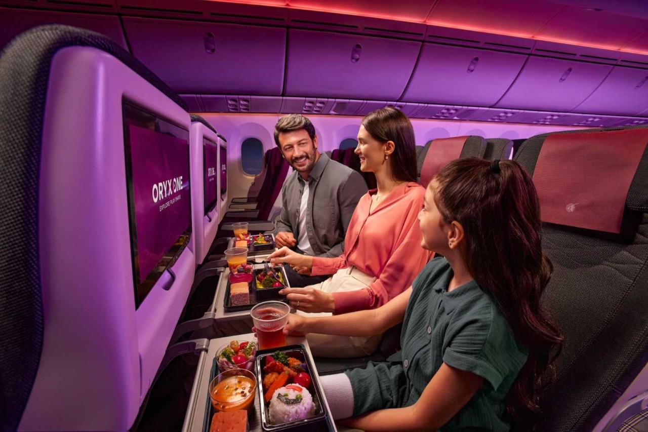 Three people in the interior of an aeroplane. They have trays of food in front of them and are laughing at each other.