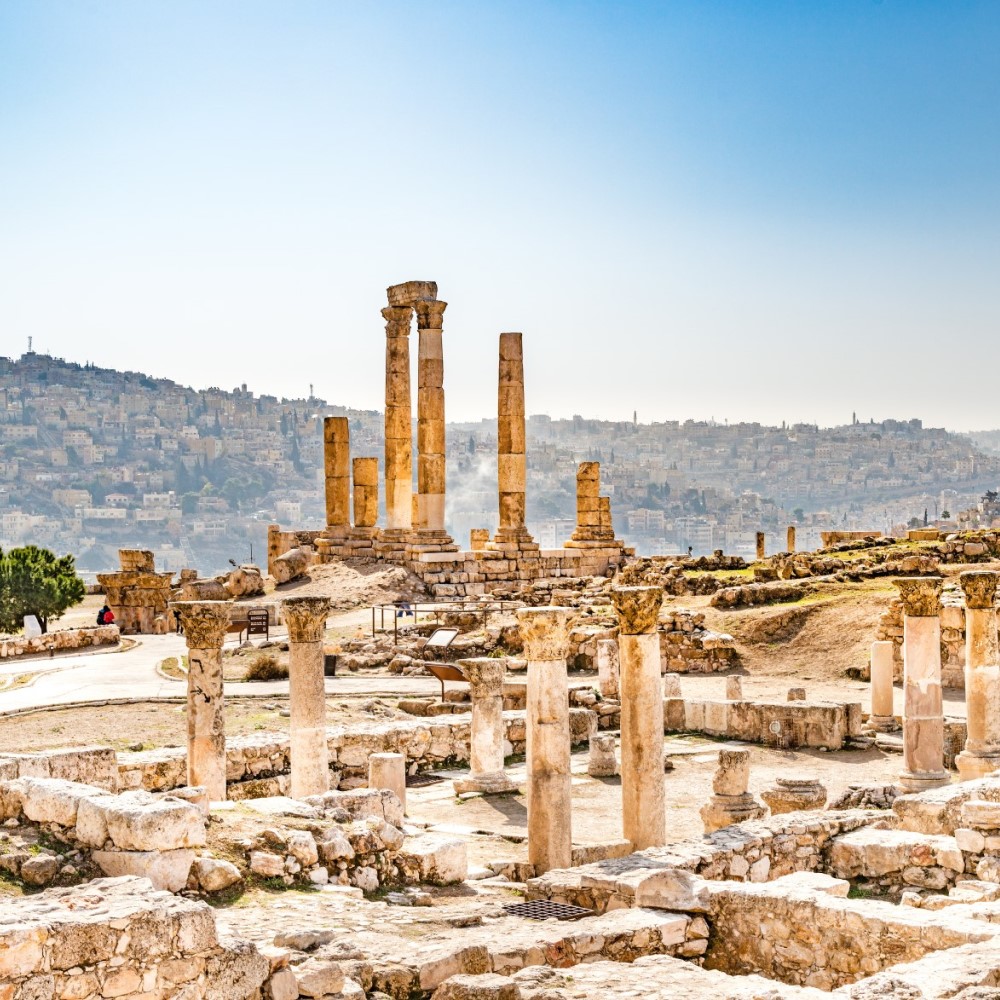 The picture shows an archaeological excavation site. The ruins and columns of a temple are prominently visible. Behind it, on two hills, extends the densely built-up city.