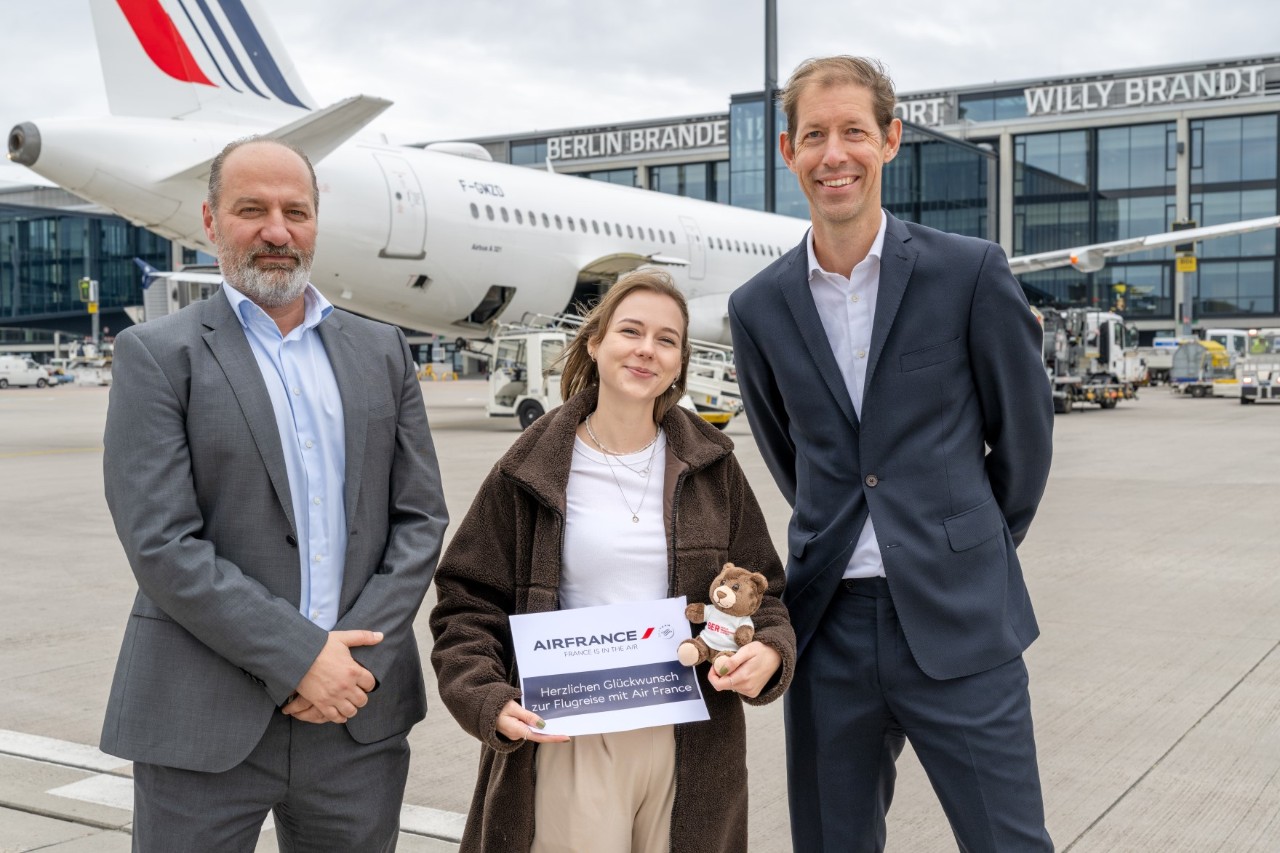 Two men and a woman pose on the apron in front of an aircraft. The woman in the middle presents a voucher and holds a teddy bear in her arms.