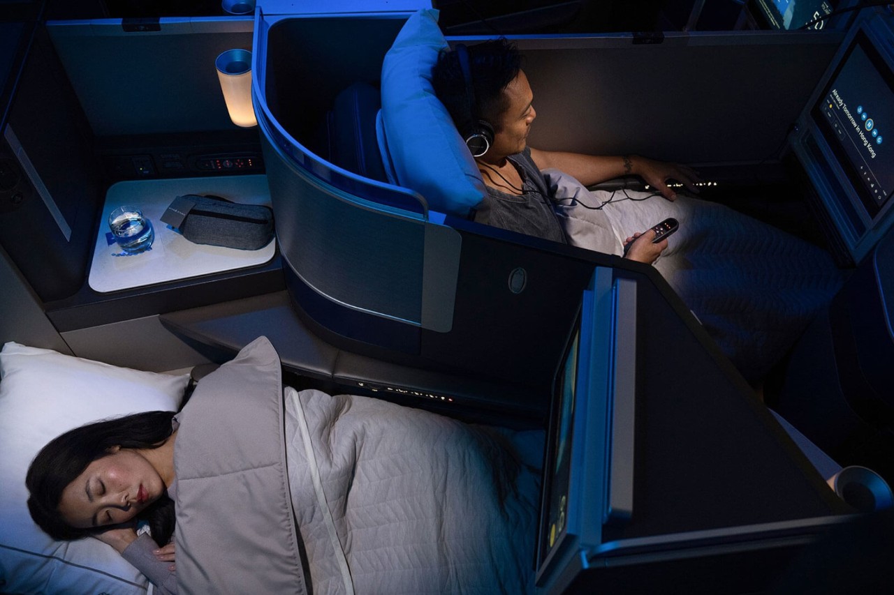 Two people sitting in comfortable aircraft seats. One is lying down asleep, the other is looking at a screen.