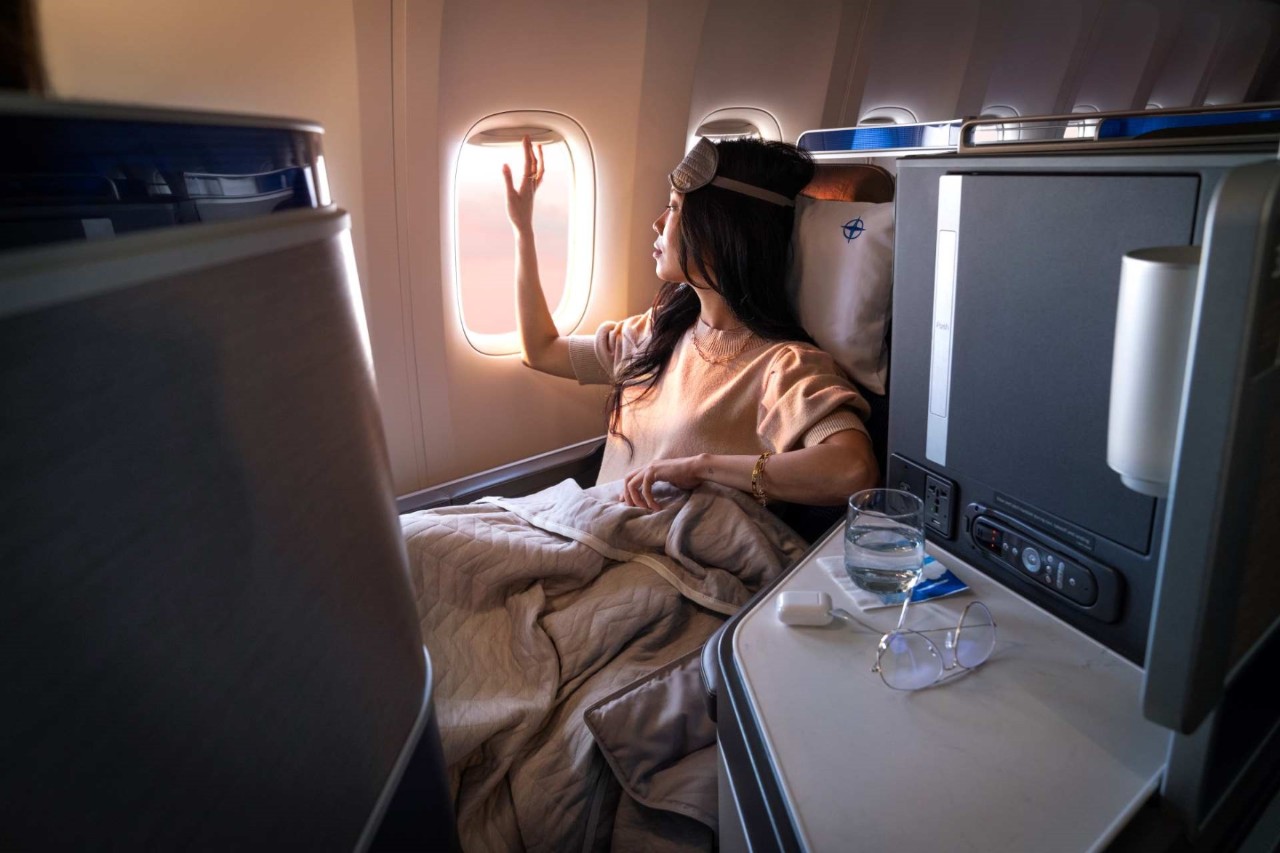 A passenger with a blanket and a sleep mask pulled up looks out of an aircraft window