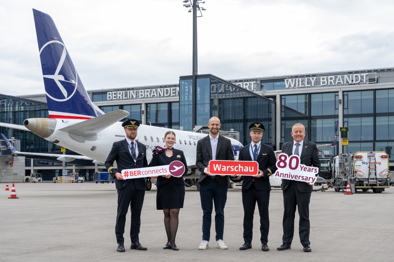 Crew members and two men in suits are posing with signs on the apron in front of an airplane.