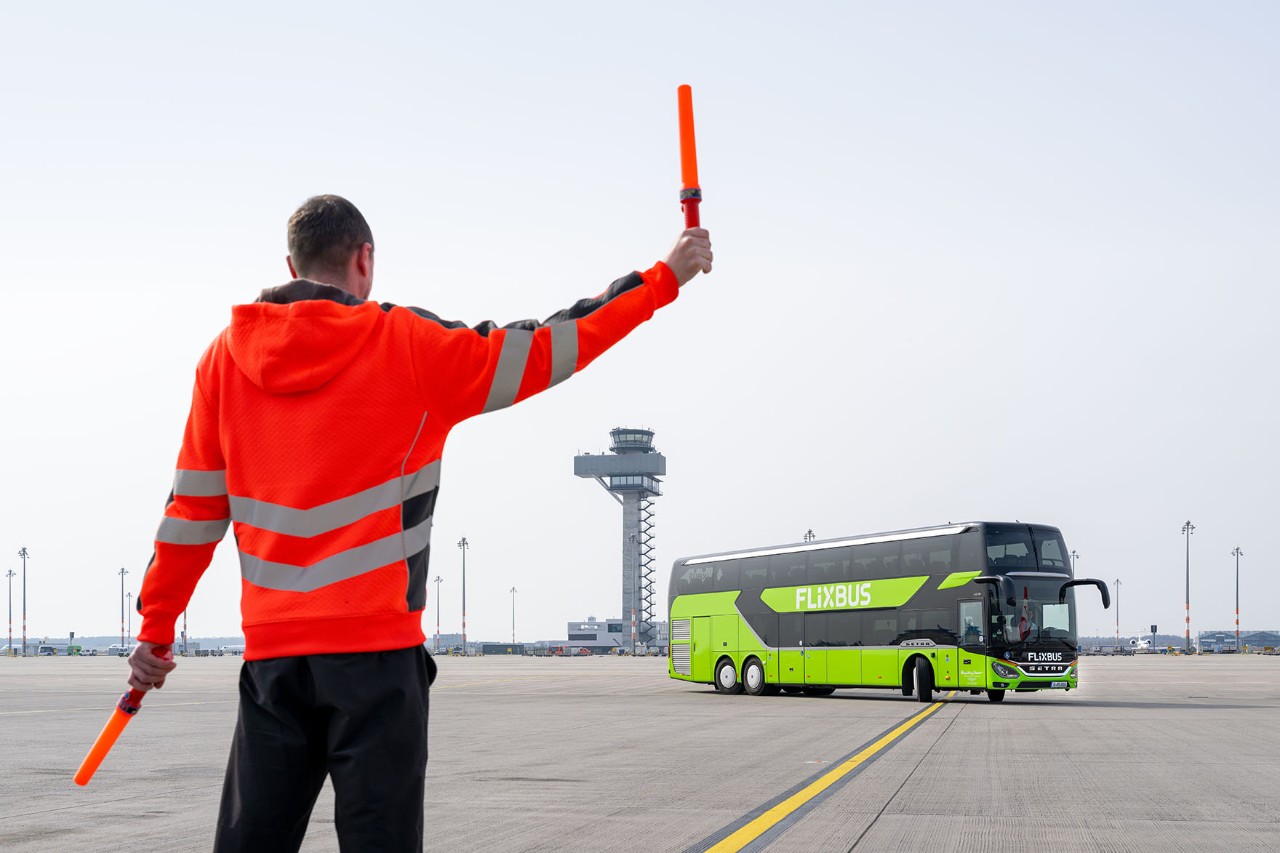 A man is standing on the apron wearing a high-visibility vest. In the background, a green long-distance coach is driving past.