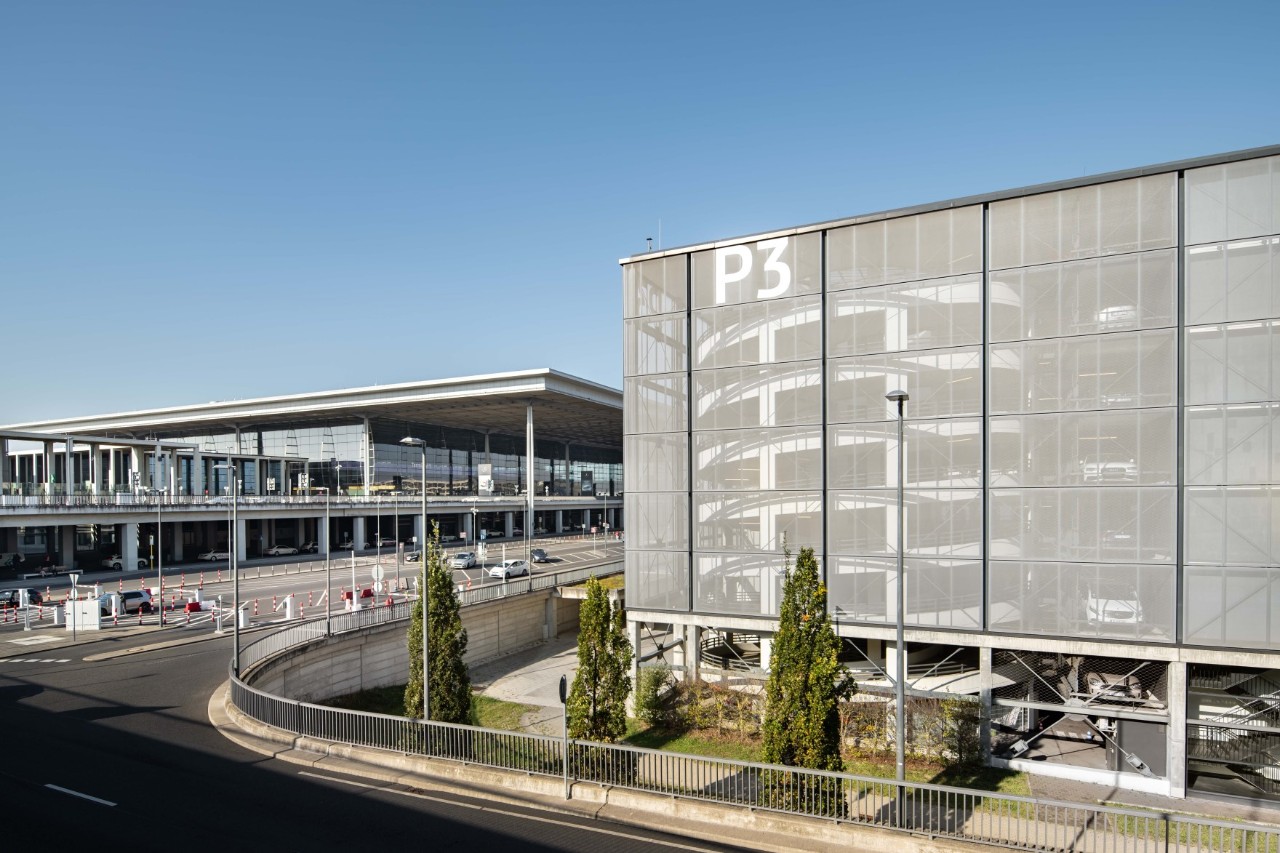 A parking garage in front on the right, Terminal 1 with its distinctive roof in the background, and a road in between.