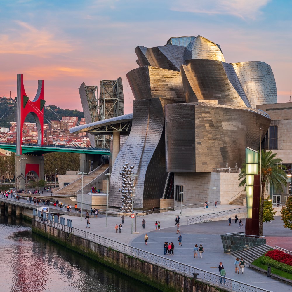 Das Bild zeigt das Guggenheim-Museum in Bilbao mit seiner geschwungenen Architektur aus Titan und Glas, umgeben von einer modernen Brücke und einer Promenade am Fluss mit Spaziergängern und Palmen.