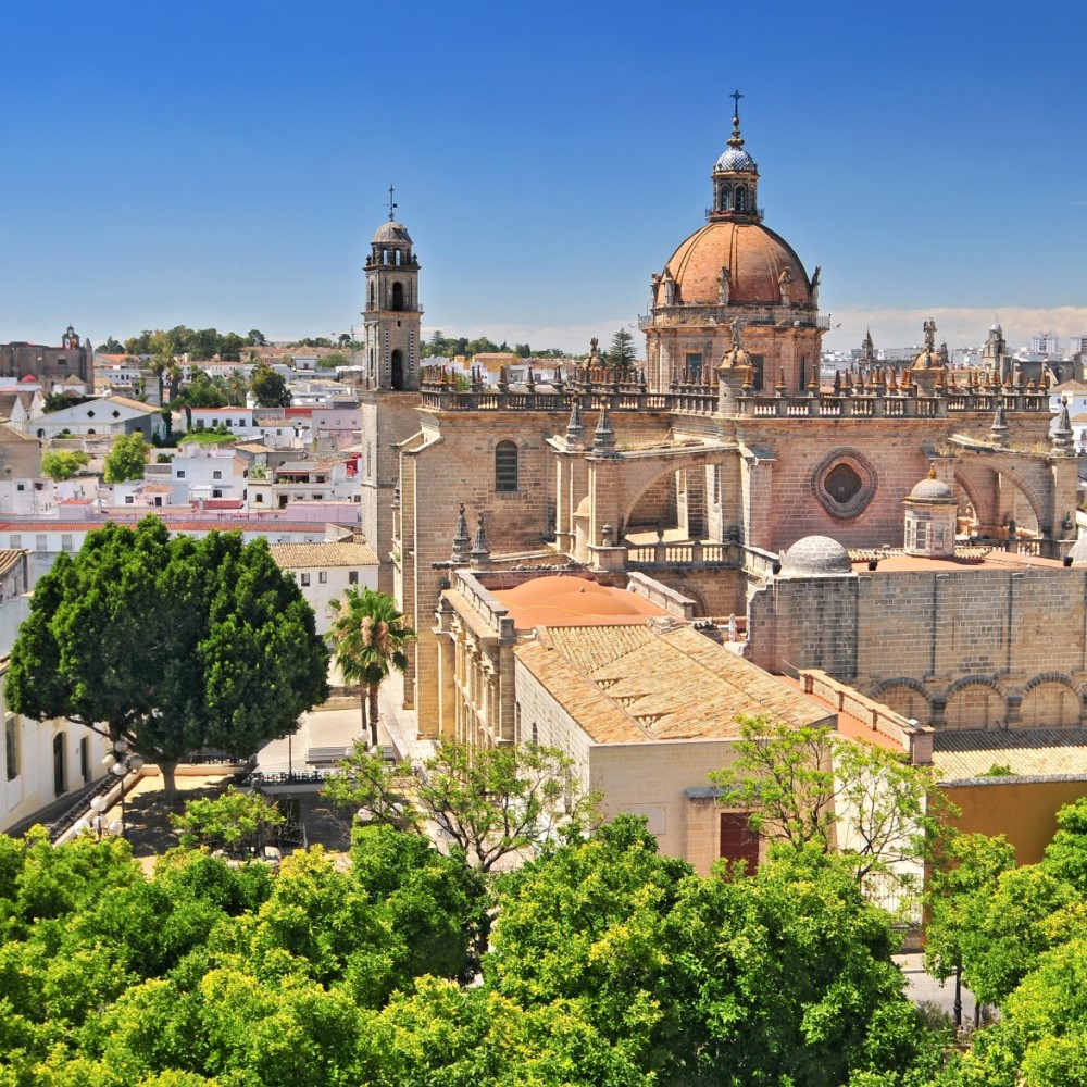 Das Bild zeigt eine gewaltige Kathedrale mit einer großen roten Kuppel, Türmen und angrenzenden Kirchengebäuden, eingebettet in eine mediterrane Stadtlandschaft. Im Vordergrund sind grüne Bäume und weiße Häuser zu sehen.