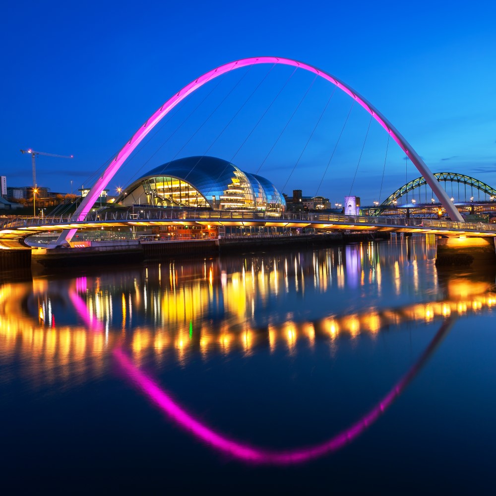 Abendliche Aufnahme eines Flusses in einer Stadt, im Vordergrund ist eine pink beleuchtete Bogenbrücke zu sehen, die sich im Wasser spiegelt. Weitere Lichter der angrenzenden Gebäude spiegeln sich im Wasser. Im Brückenbogen ist ein modernes Gebäude sichtbar, im Hintergrund eine zusätzliche Bogenbrücke. Rechts im Bildvordergrund steht direkt am Uferrand ein weiteres beleuchtetes Gebäude.