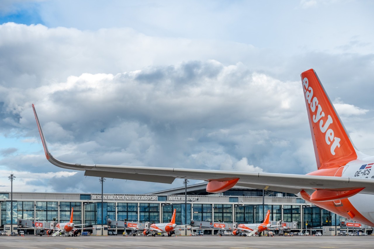 Blick unter dem Flügel eines easyJet-Flugzeugs hindurch auf das Terminal-Gebäude des BER, vor dem weitere easyJet-Maschinen auf Abfertigungspositionen stehen